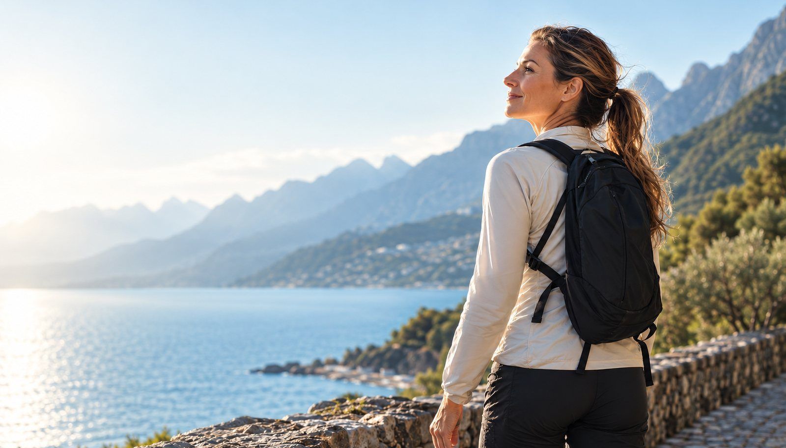 Walker with a backpack following a stone lakeside path towards sunlit mountains